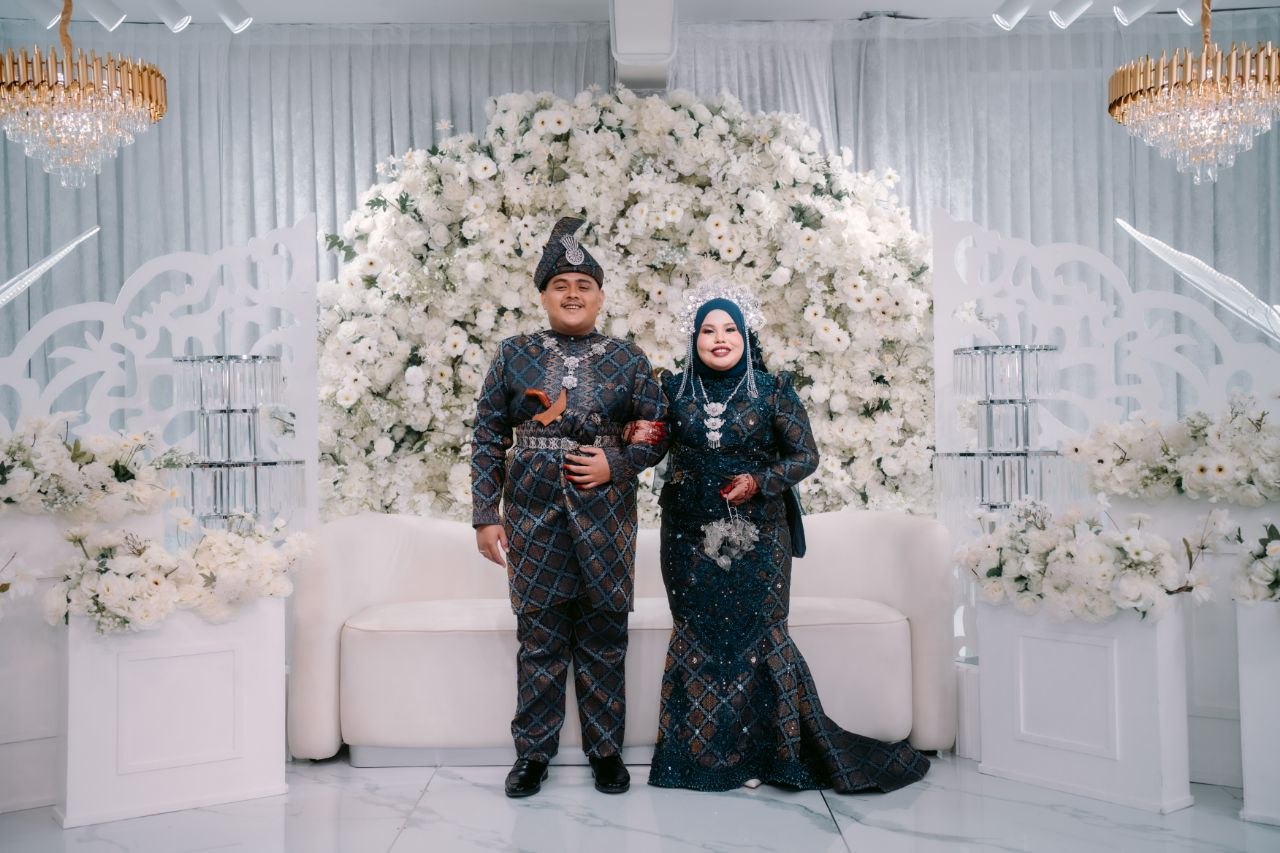 Aboy dan Emylia berpakaian biru gelap di ruang majlis berhias bunga putih
